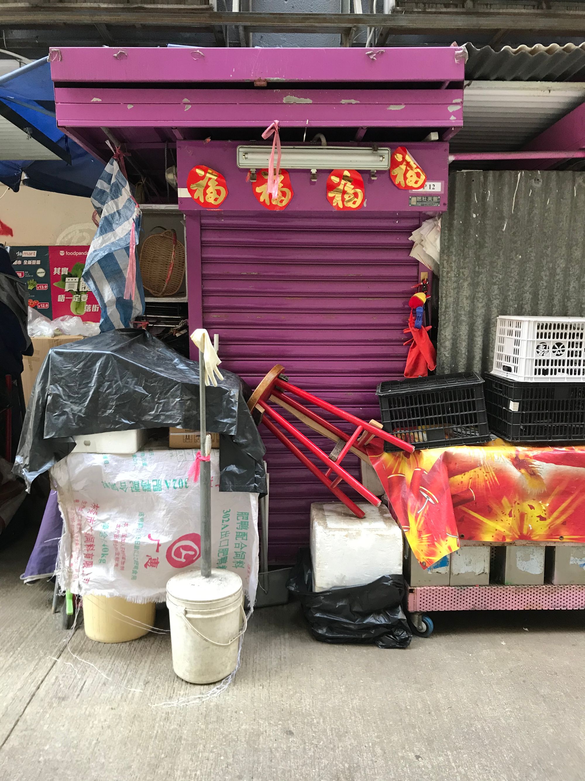 A closed street stall cabinet painted in purple with boxes  stacked in front of it and a red stool titled at an angle.