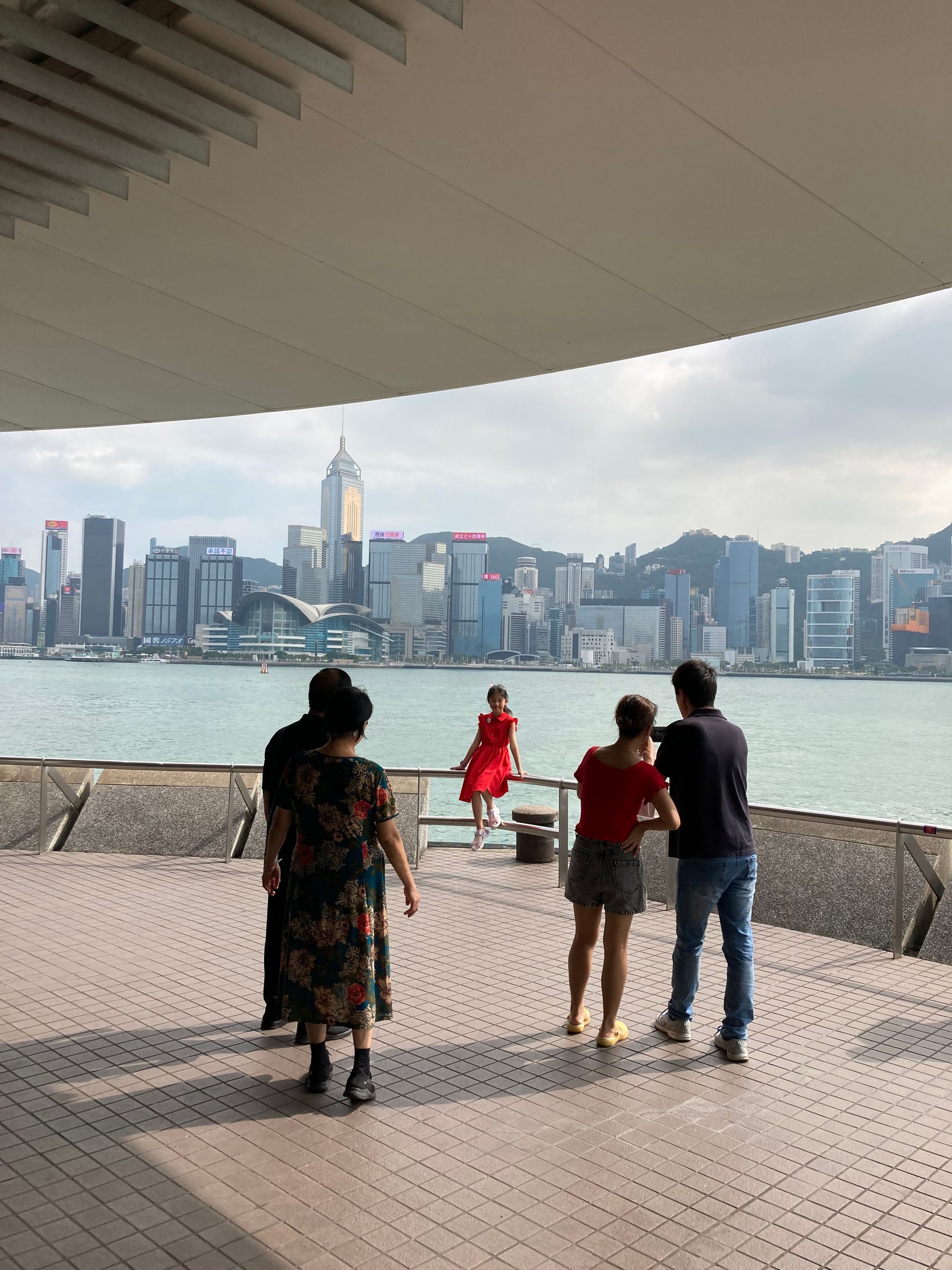 A girl in red poses before the HK skyline while her parents take a photo of her. Her grandparents are on the left watching her.