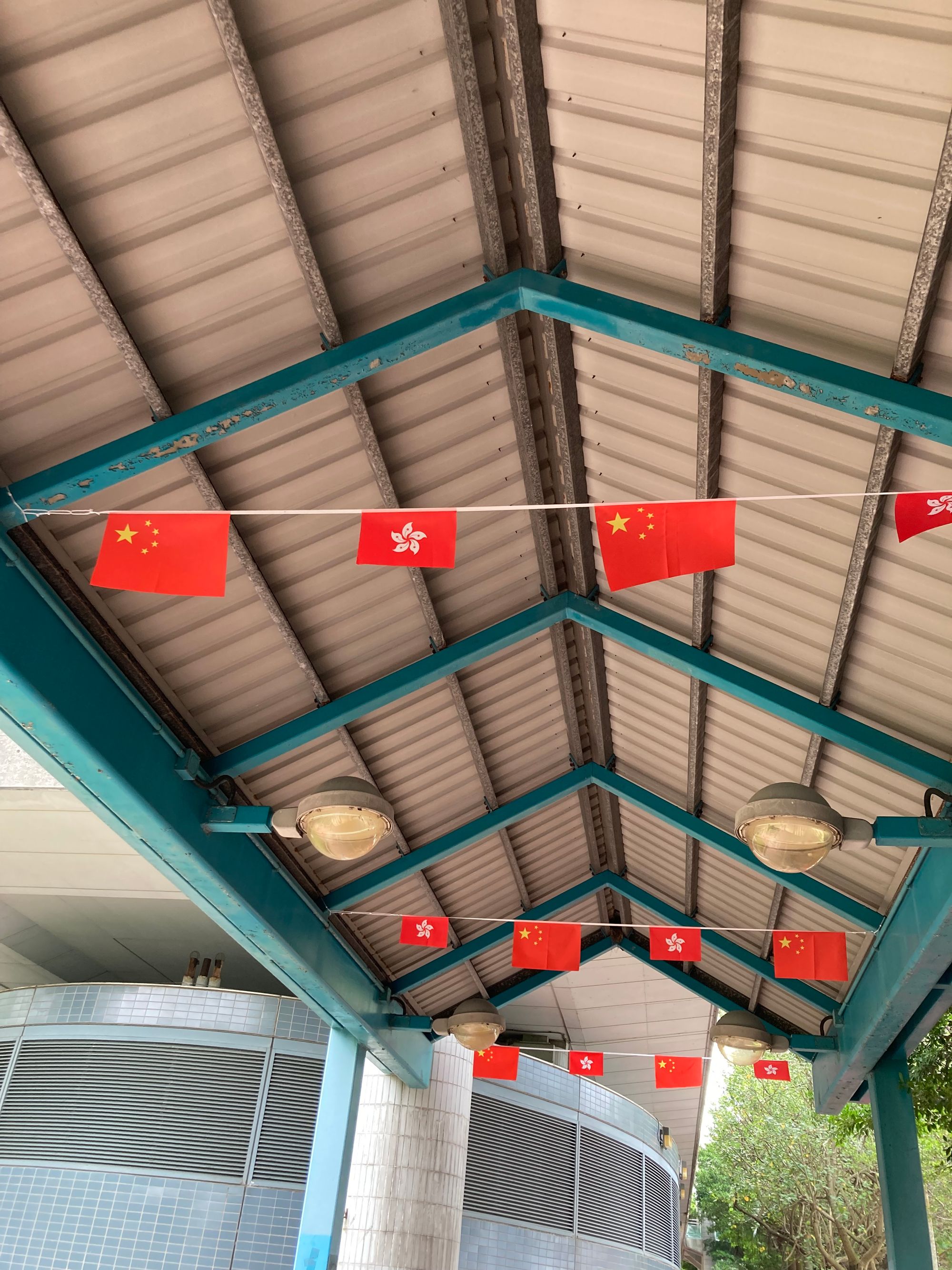 A string of mainland and HK flags decorates the top of a public walkway. The HK flag is noticeably smaller than the mainland one