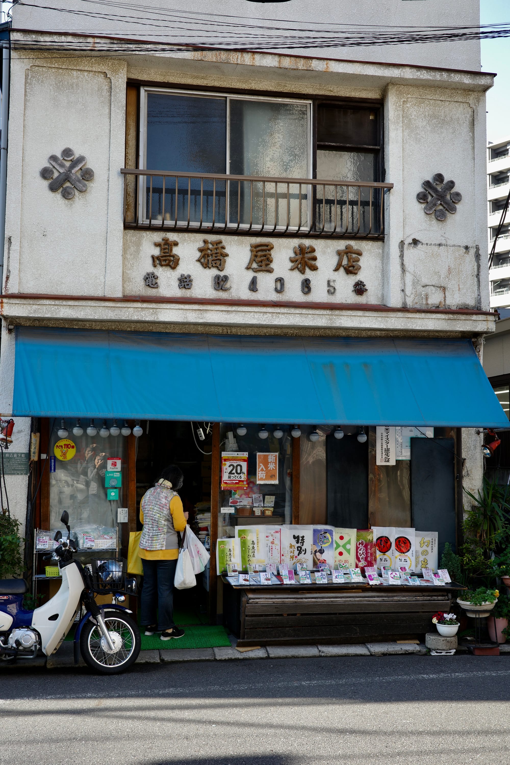 A two-storey rice store with colourful packets of rice displayed outside. A woman lingers by the doorway with plastic bags on her arm. 