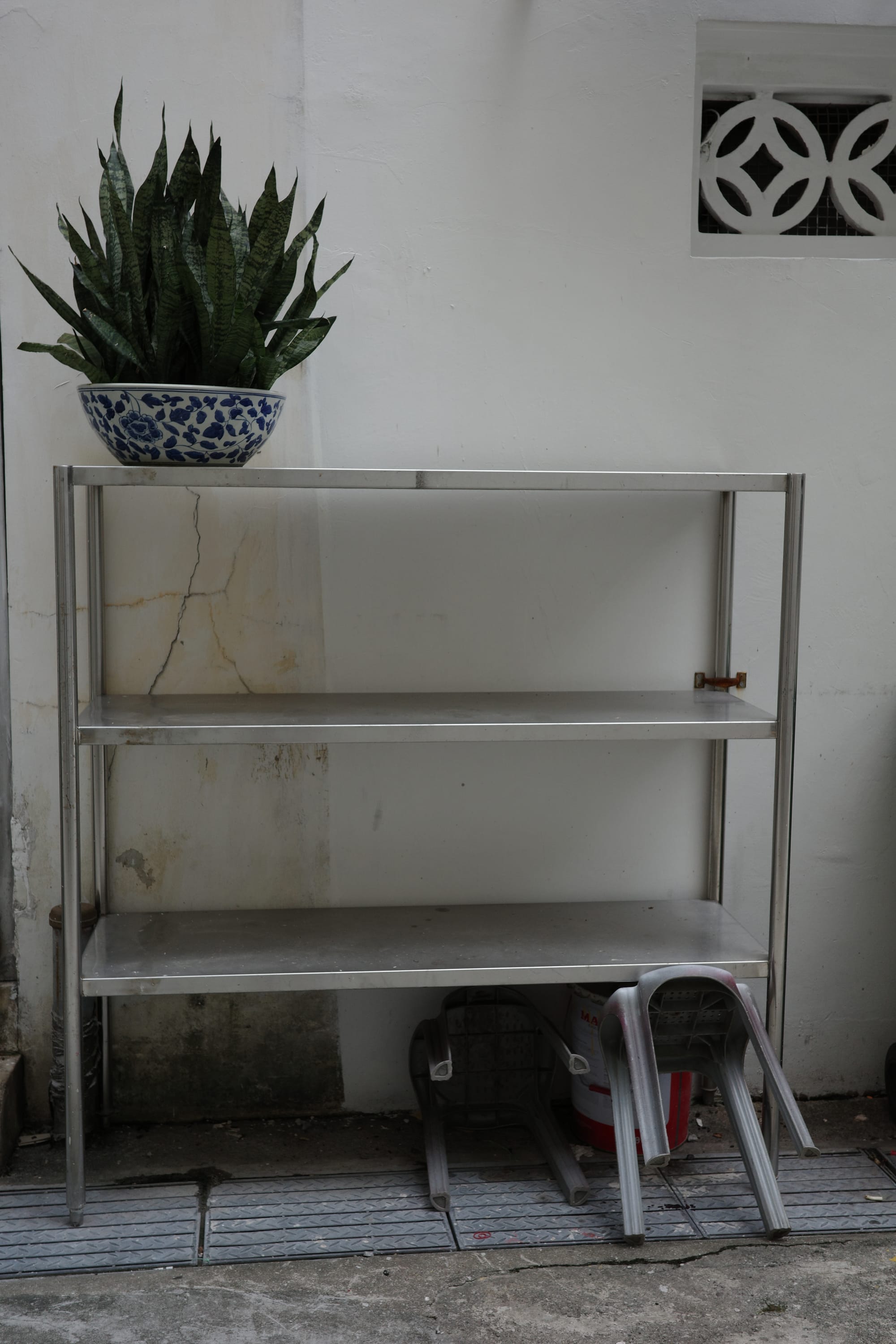 An open metal shelf against the wall. There is a potted plant placed on the left of the top shelf. Two stools are tucked under the bottom shelf on the right.