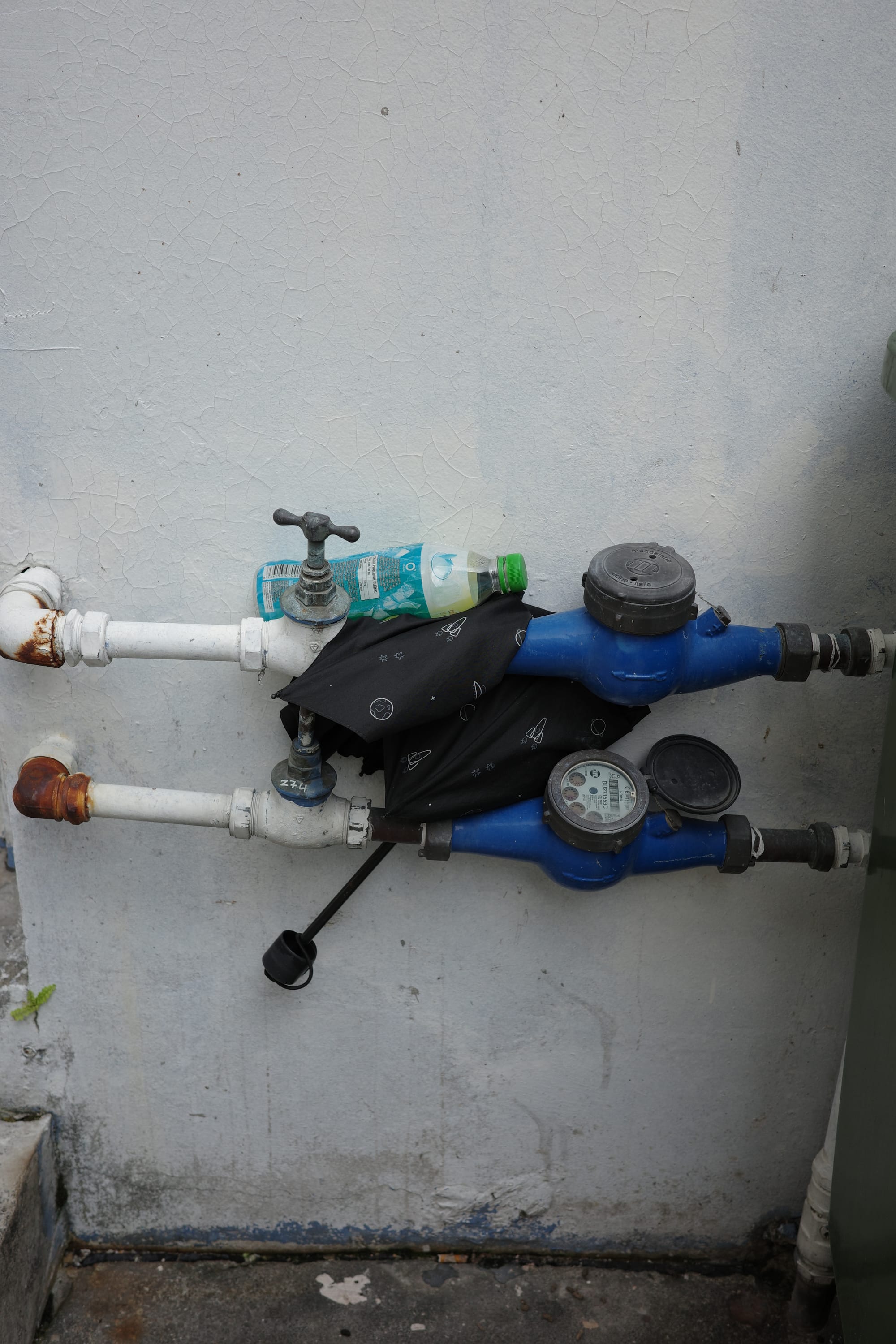 An umbrella and a plastic bottle are tucked behind some pipes and water meters in a back alley