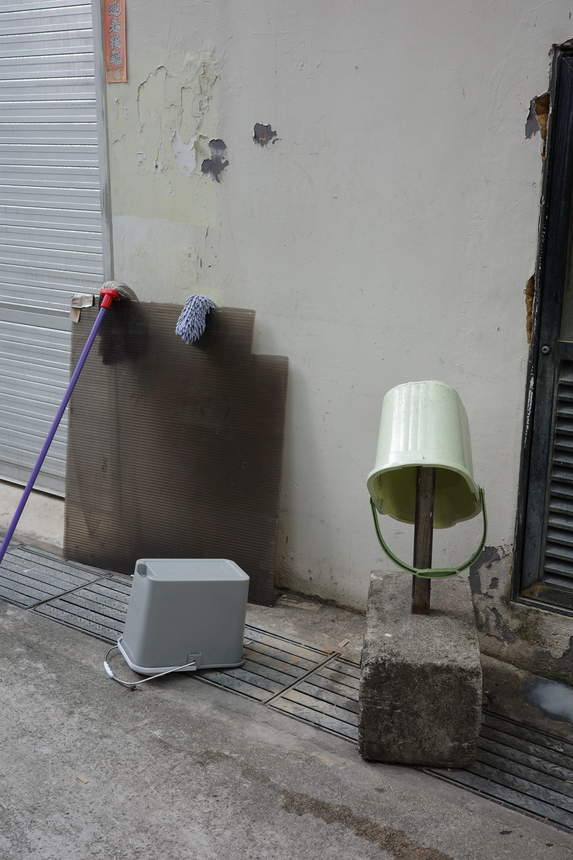 Two mops have been propped against a random sheet of hard plastic to dry. A grey mop pail sits upside down. A green mop pail has been turned over to dry on metal stump with a concrete ballast.