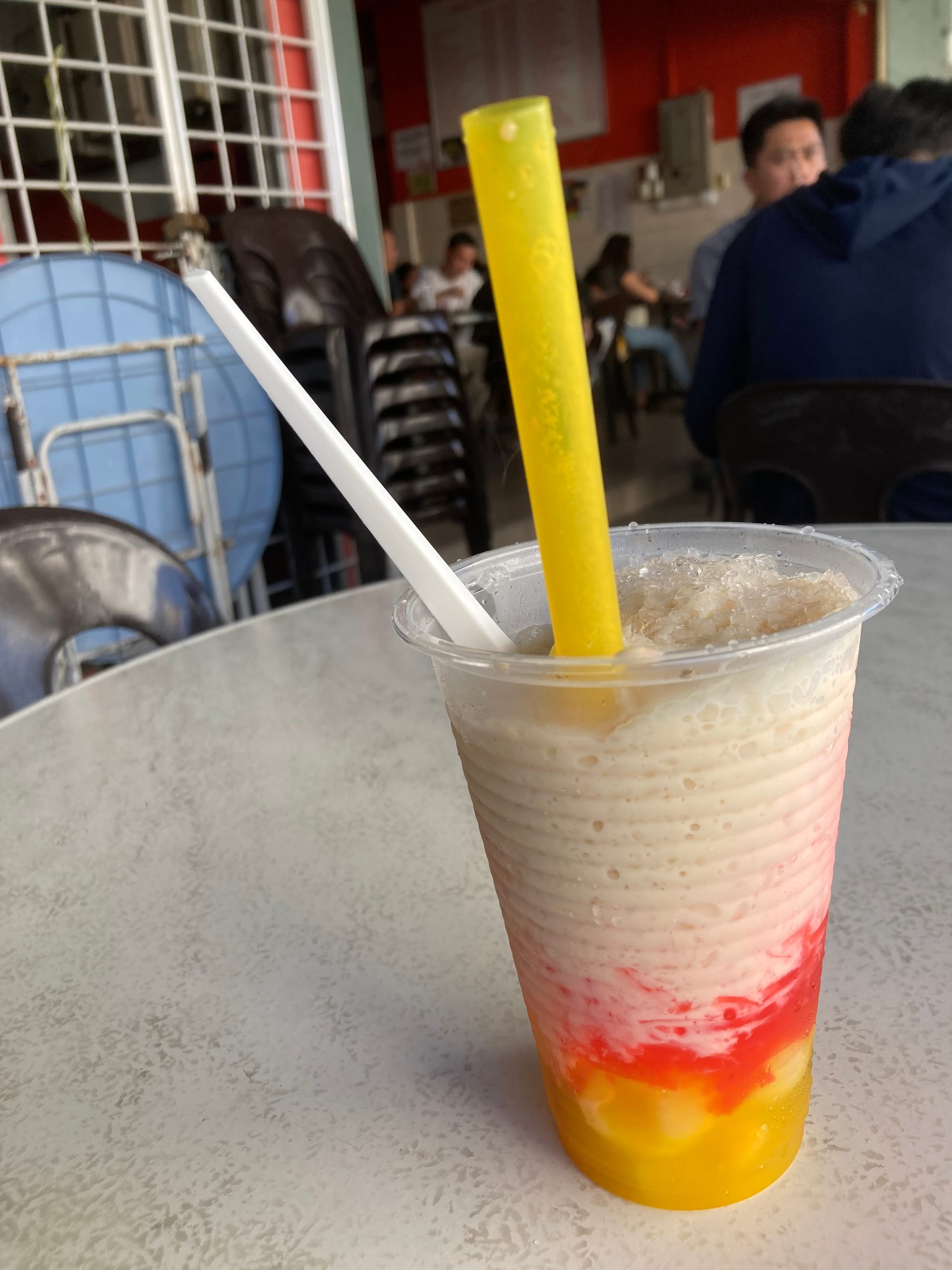 In the foreground is a plastic cup with yellow straw holding a drink that is yellow at the bottom, then red, then pink and then white with shaved ice at the top. 