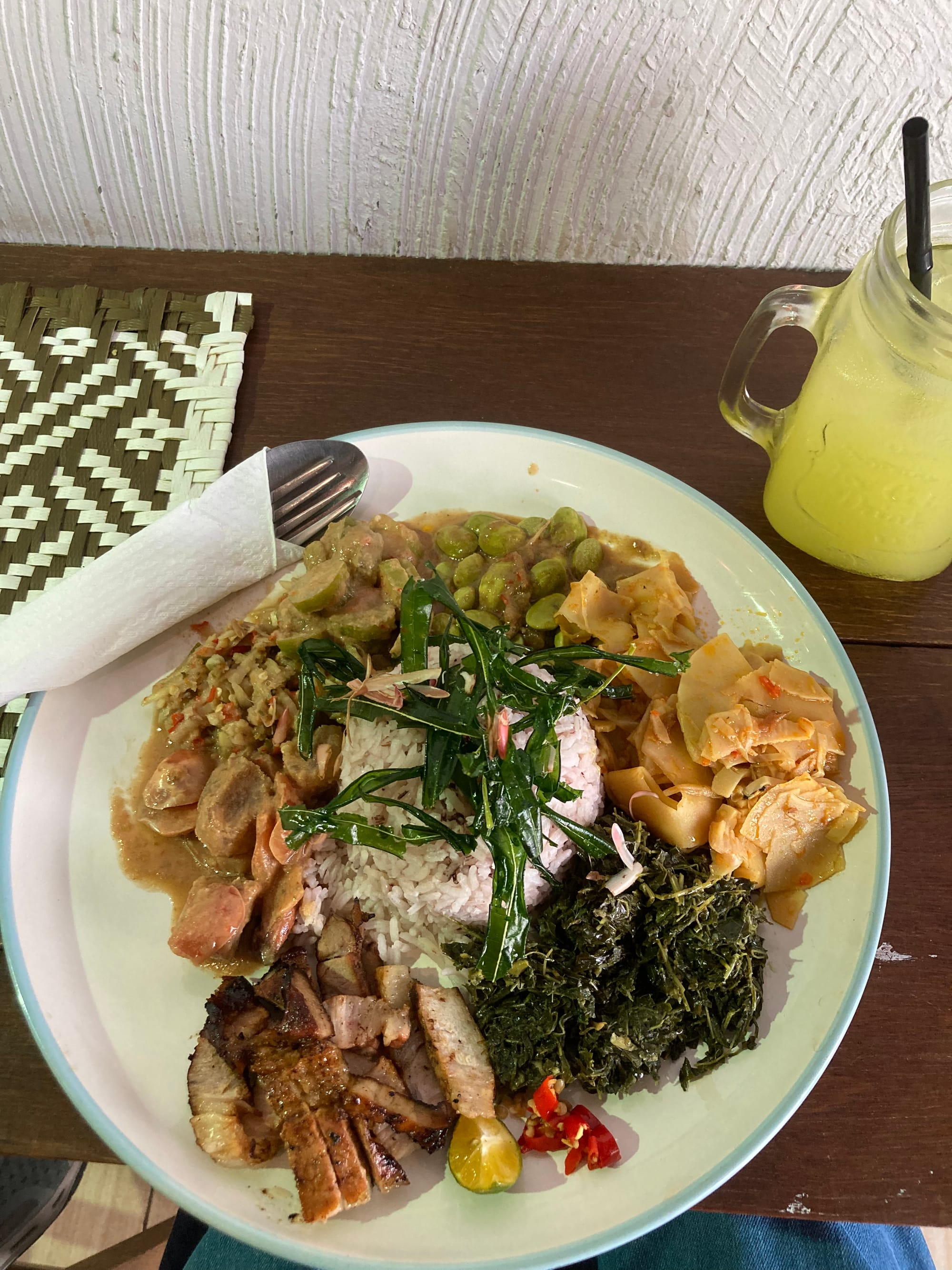 A plate of rice encircled by a variety of vegetables, some roast pork, chilli, and lime