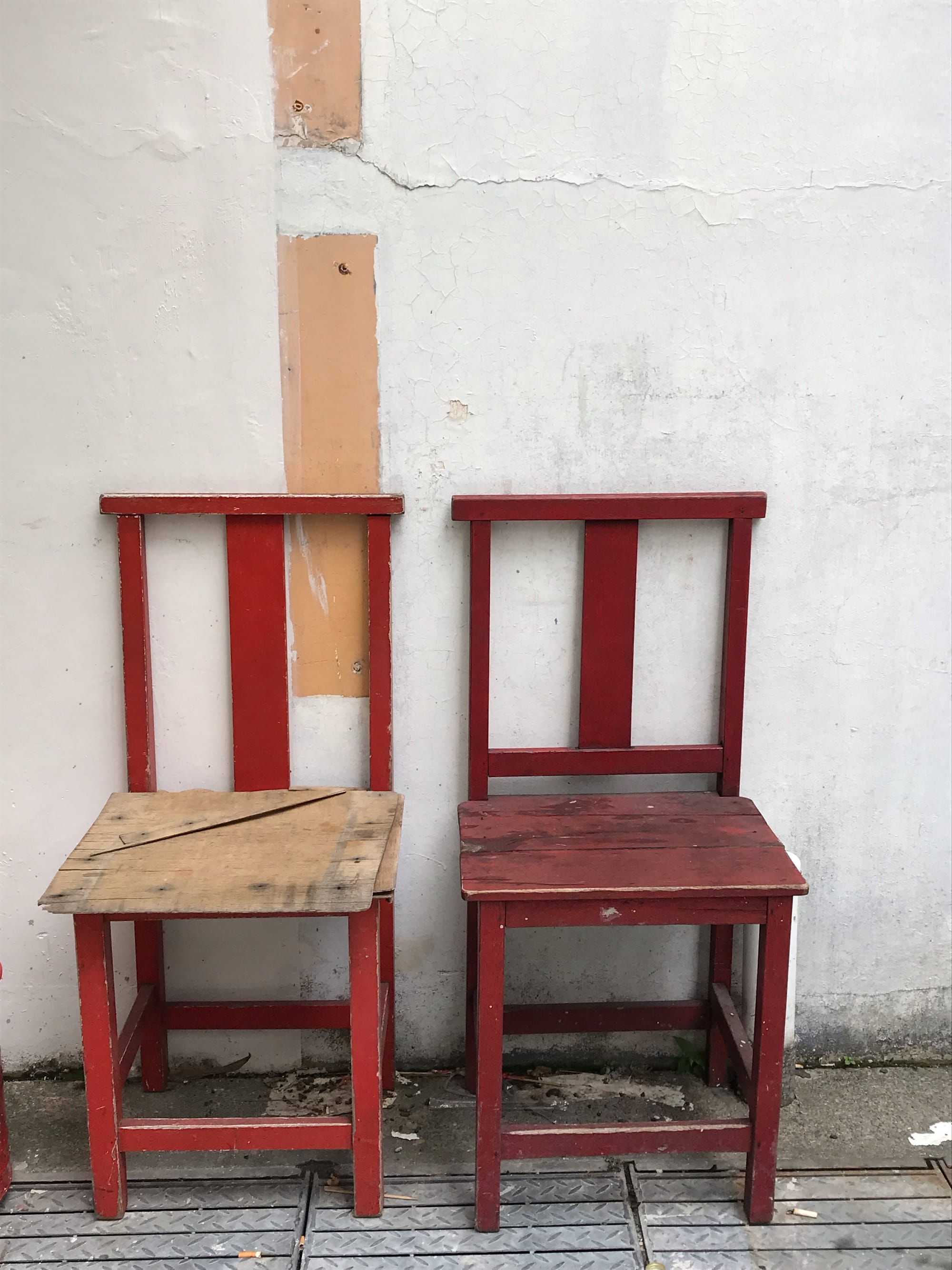 Two worn, red-stained wooden chairs sit next to each other in an alley