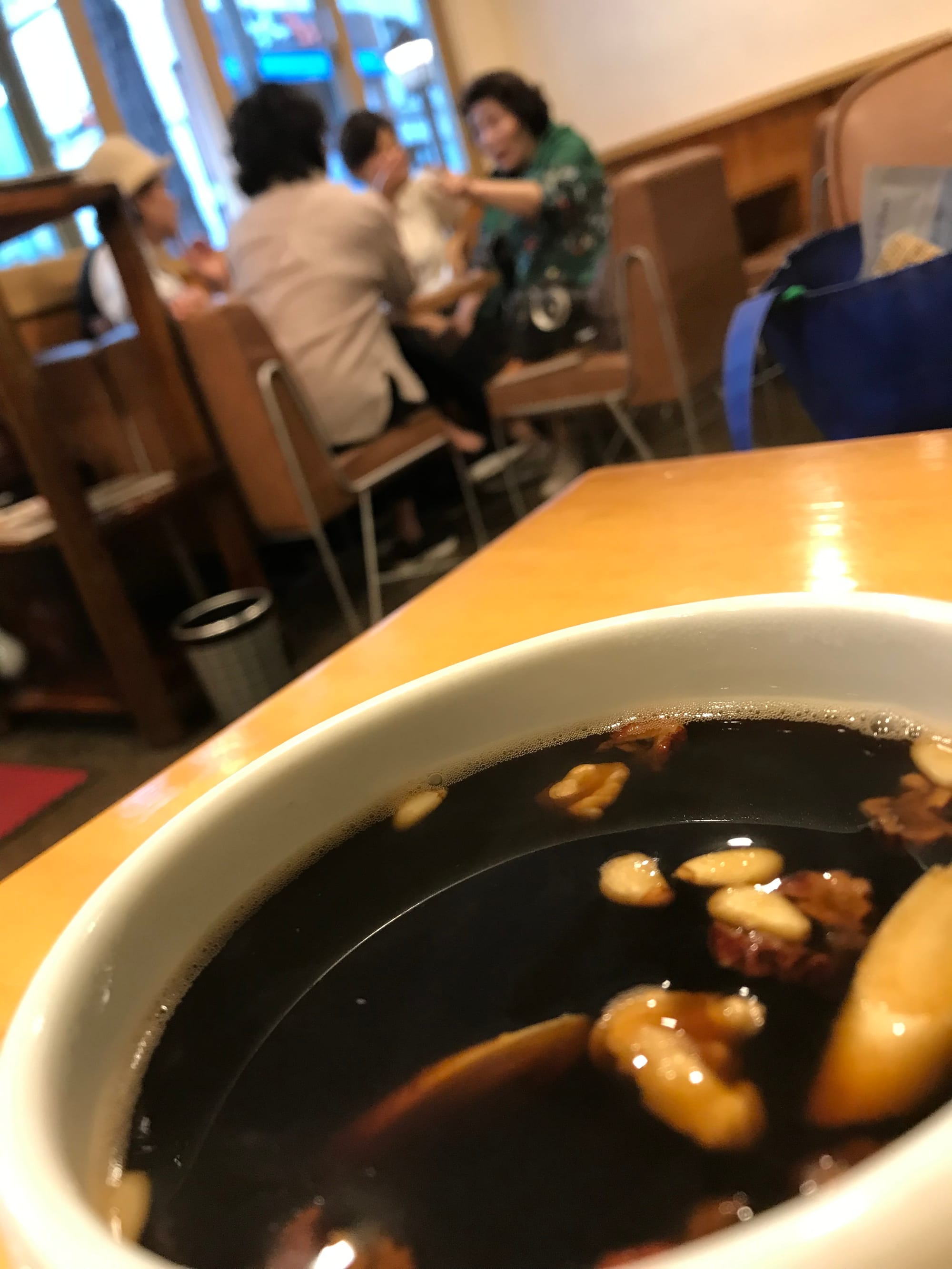 in the foreground is a bowl of a medicinal herbal brew. In the background is a blurry group of women in an animated conversation.