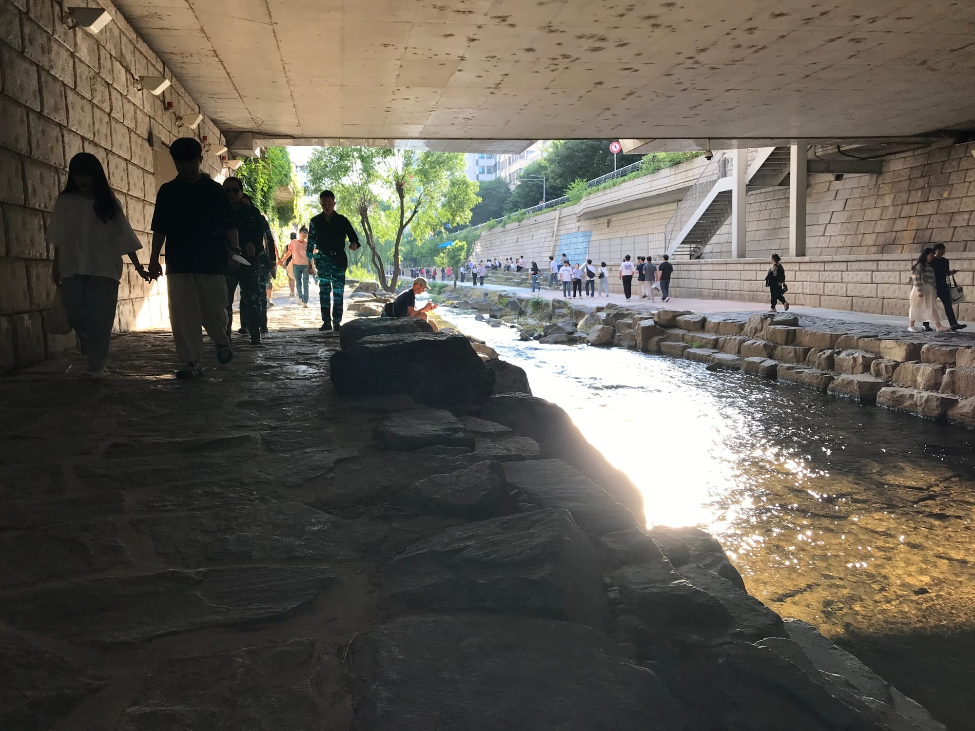 People are walking by a stream beneath an underpass. The sun glints off the water.