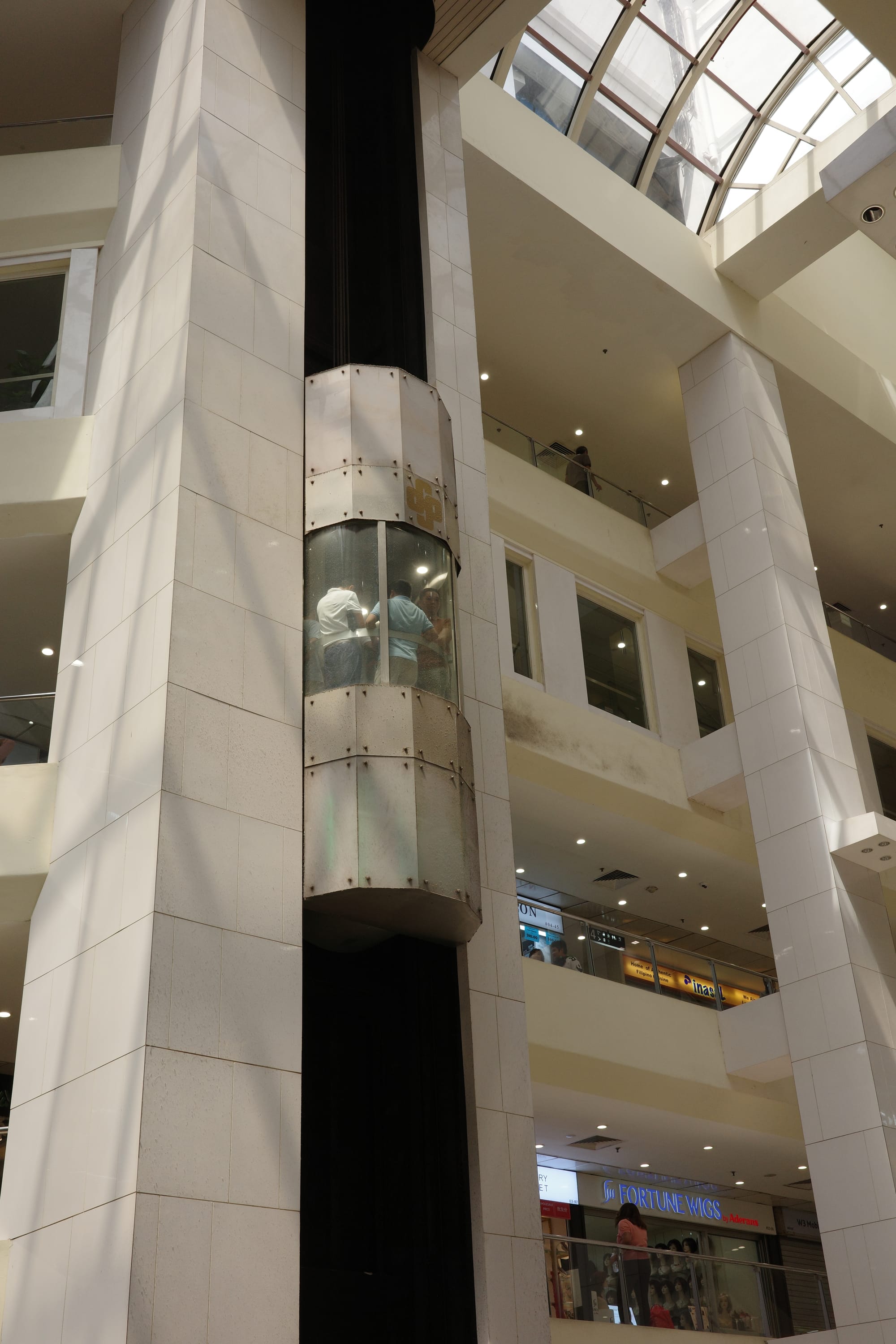 A lift with a window in it stops at a floor in a mall. There are two men in the lift.