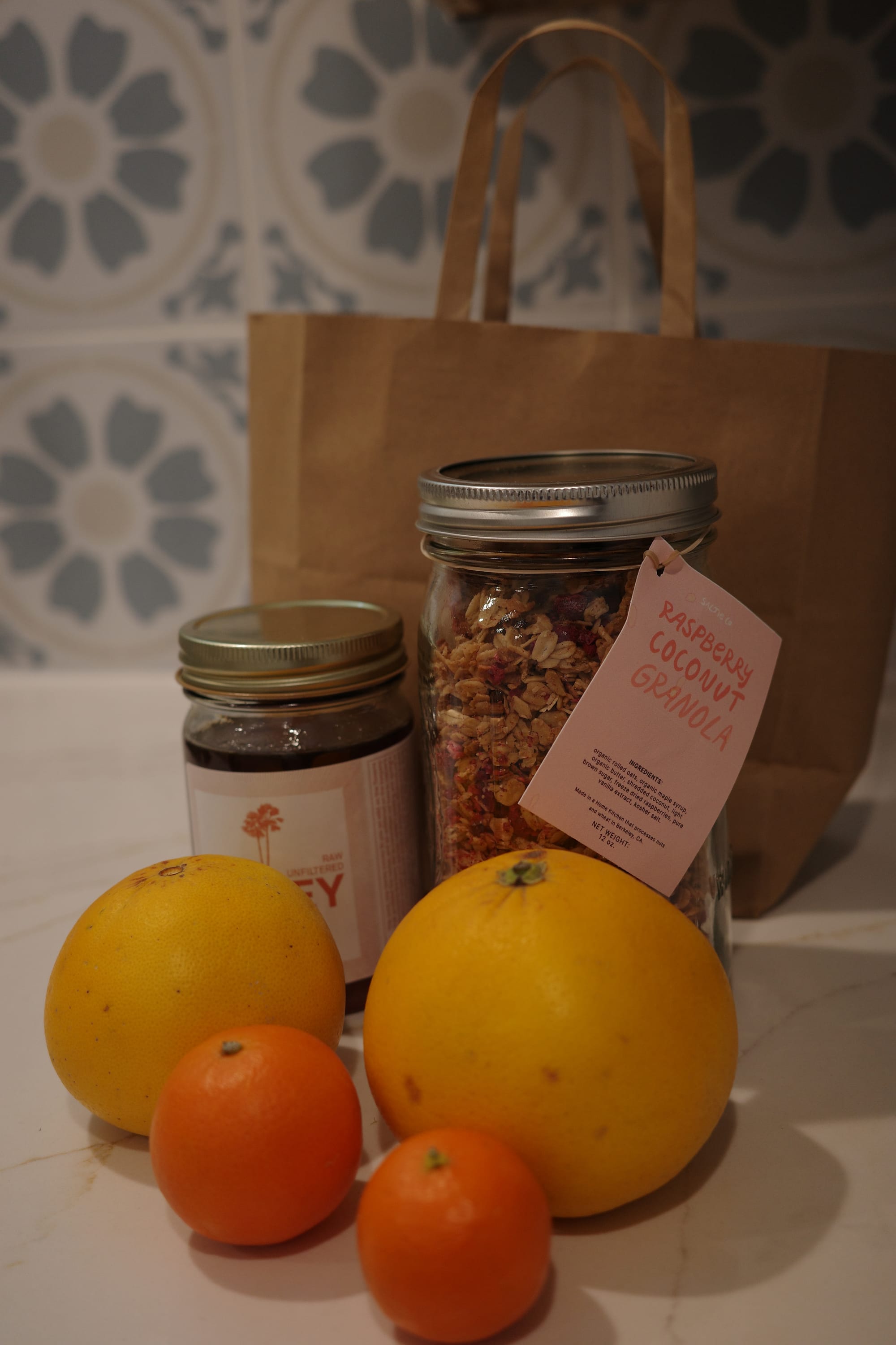 A bottle of granola stands next to a shorter bottle of honey. There are two grapefruits and two small citrus fruits in front of the bottles.