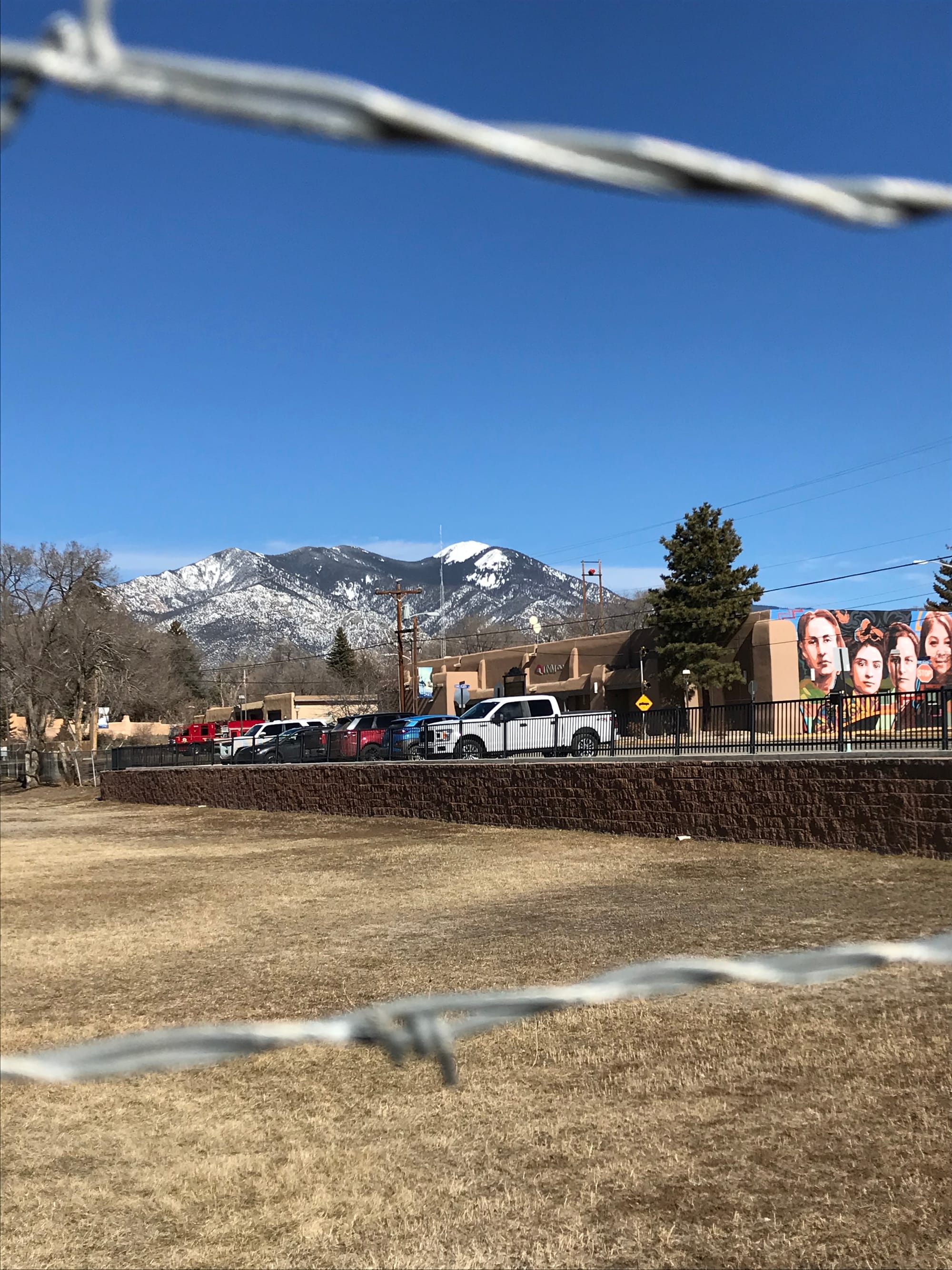 In the foreground, two barbed wires lines; in the middle, a lot with short dry grass; a parking lot just above with a street-art mural depicting young New Mexicans; and, in the background, Taos Mountain, with the image of George Washington.