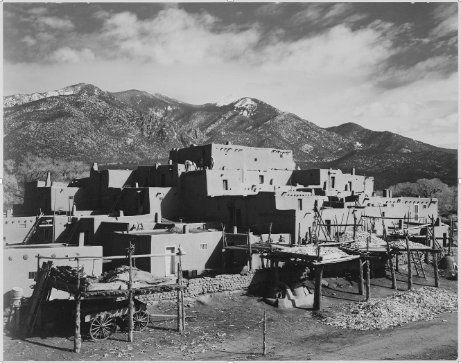 A black-and-white photograph (taken in 1941 by Ansel Adams) of the north houses at Taos Pueblo with Taos Mountain and an image of George Washington on one of its peaks.