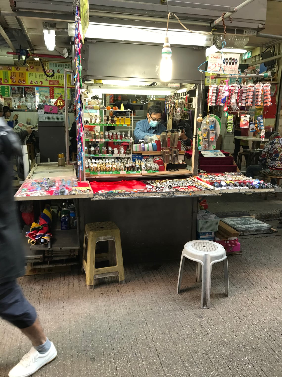 A seller of watches sits within a street stall cabinet. There are displays of watches and watch batteries on sale.