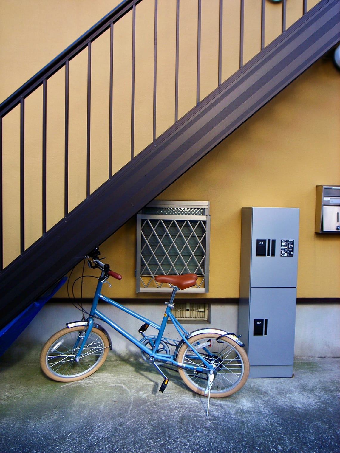 A blue bicycle sits beneath a flight of stairs, which cuts a khaki wall in a dramatic diagonal.