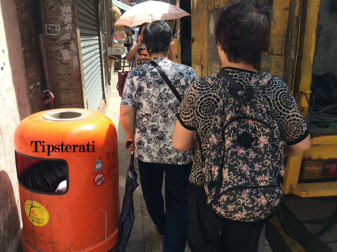 Two older ladies in boldly patterned tops walk next to an orange rubbish bin on the street.