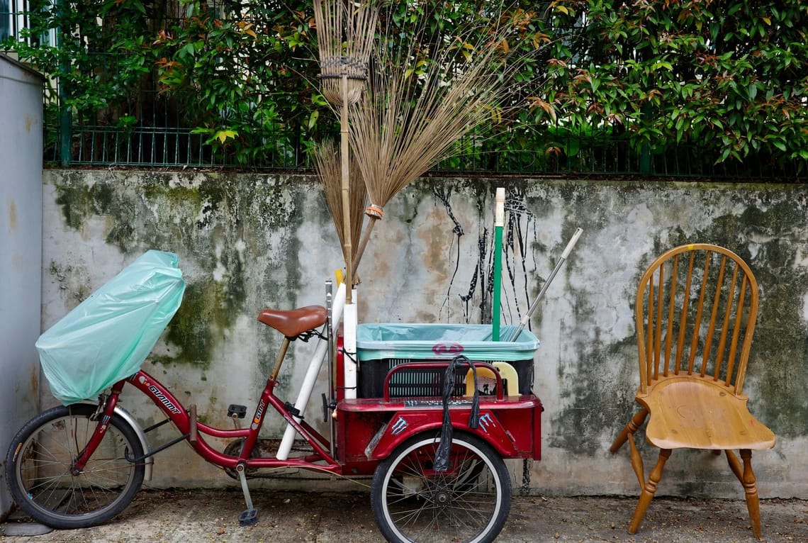 A red, adult tricycle with brooms protruding in the storage section at its back is parked next to a wooden chair