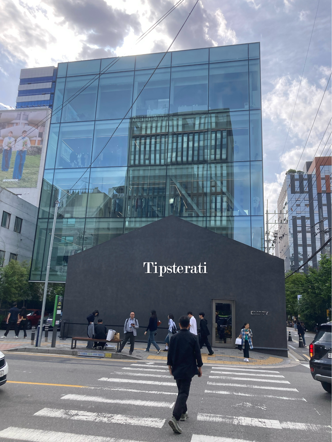 A man crosses the street in front of a glass building that rises up behind a small grey house