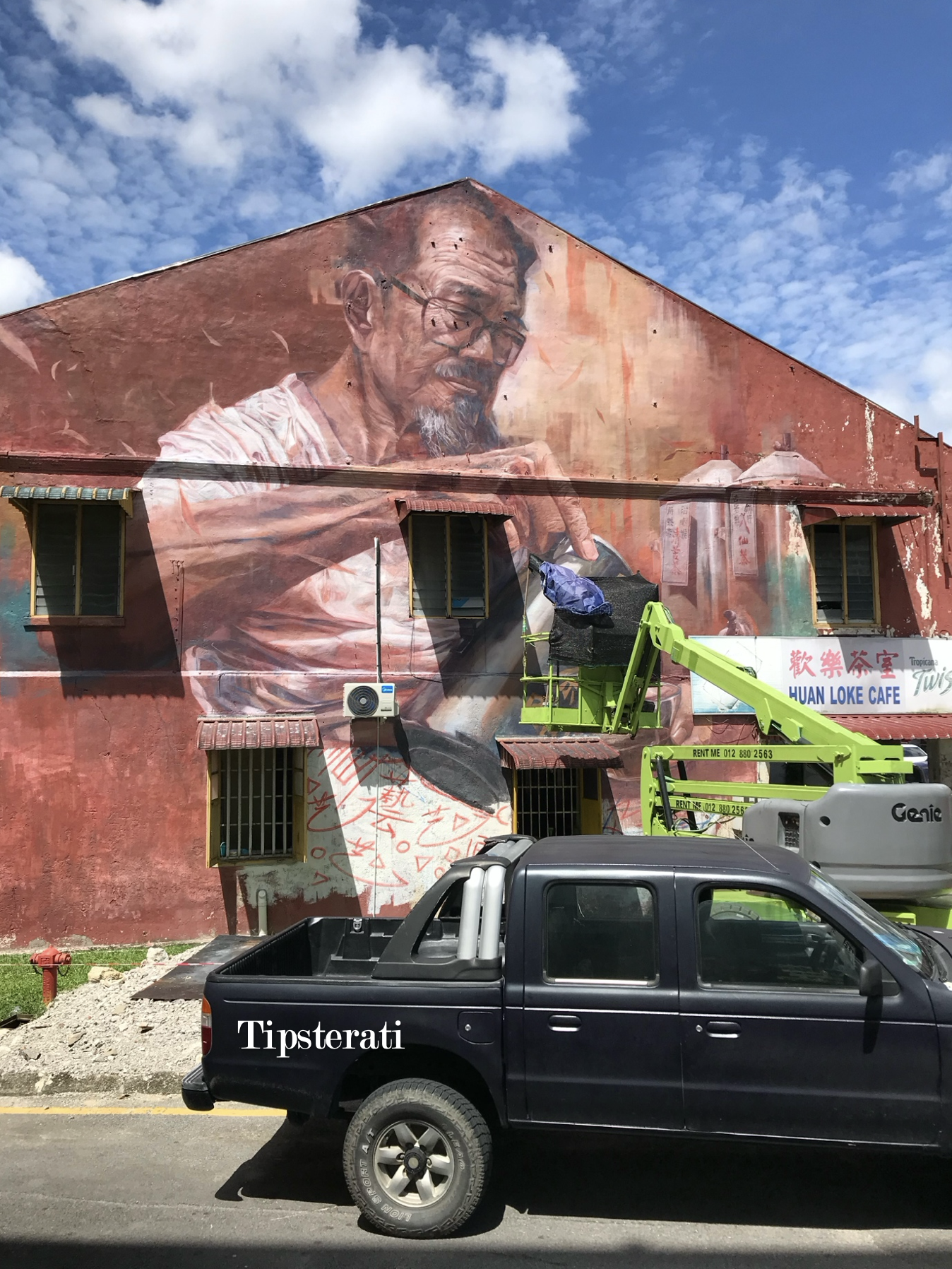 An unfinished mural on a wall with a green boom lift and a black truck in front of it.