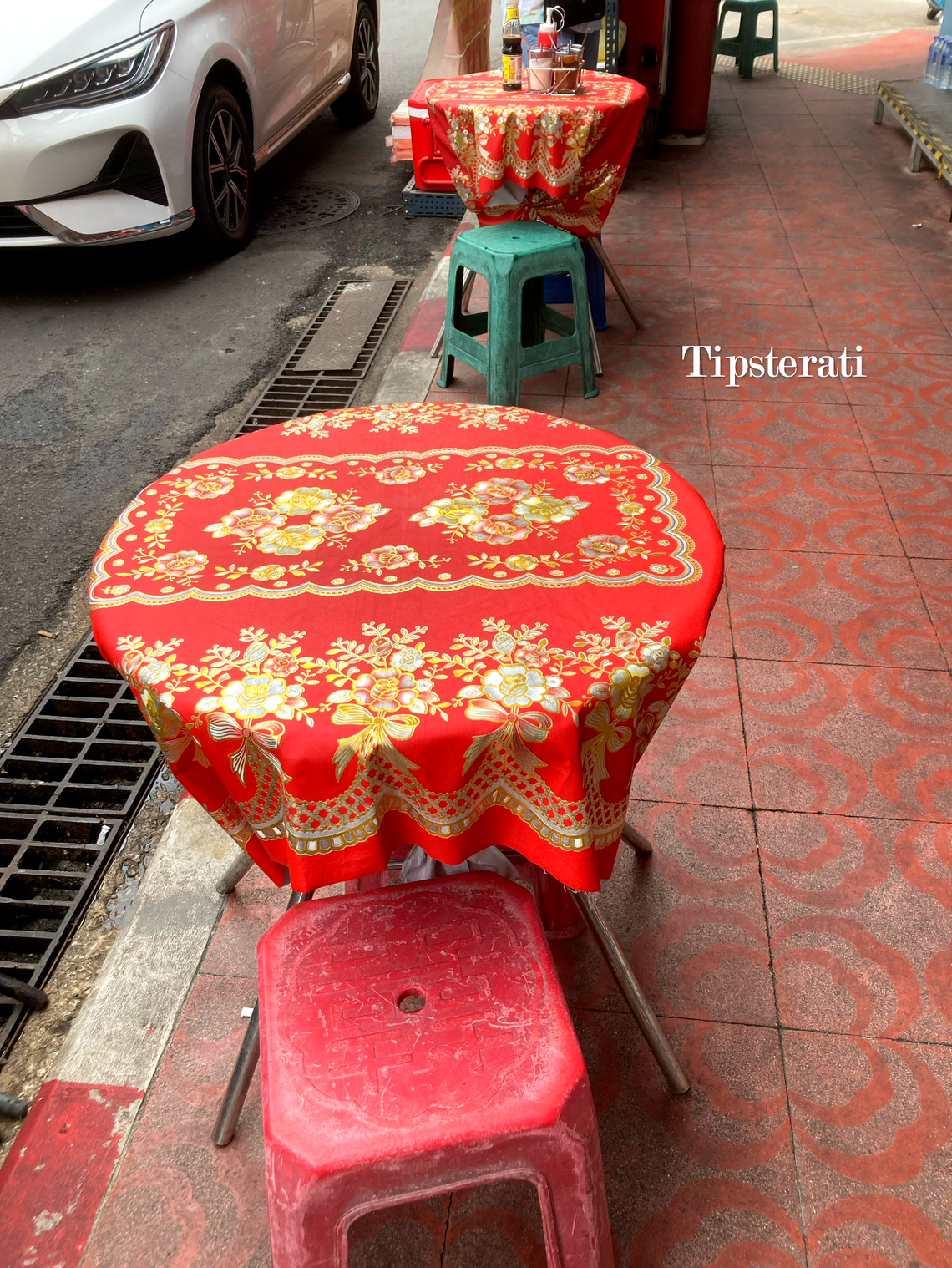 A table covered in a red patterned tablecloth with a red plastic stool on a sidewalk